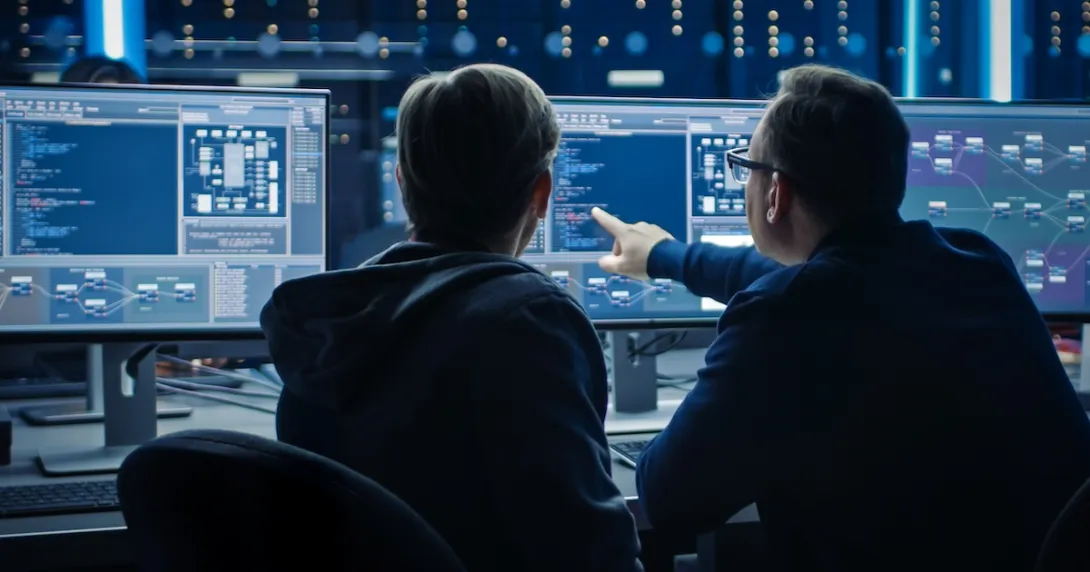 Two people sitting in front of two computer monitors looking at data on the screens Two people sitting in front of two computer monitors looking at data on the screens