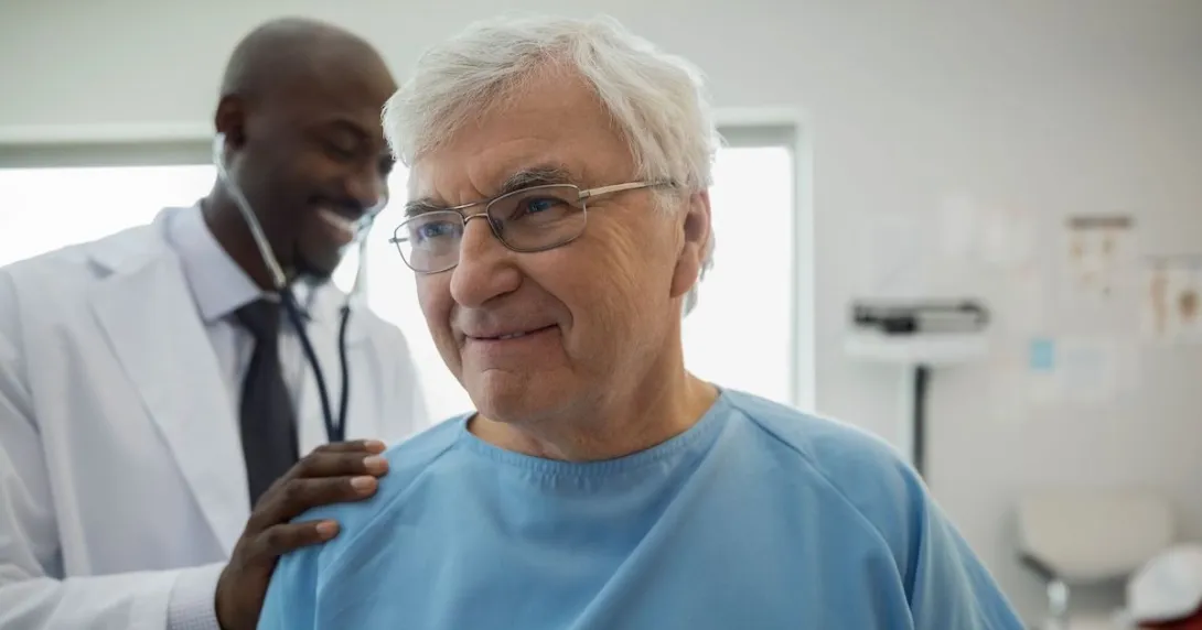 
  Healthcare provider in a lab coat standing behind a patient in a blue hospital gown checking their vitals
