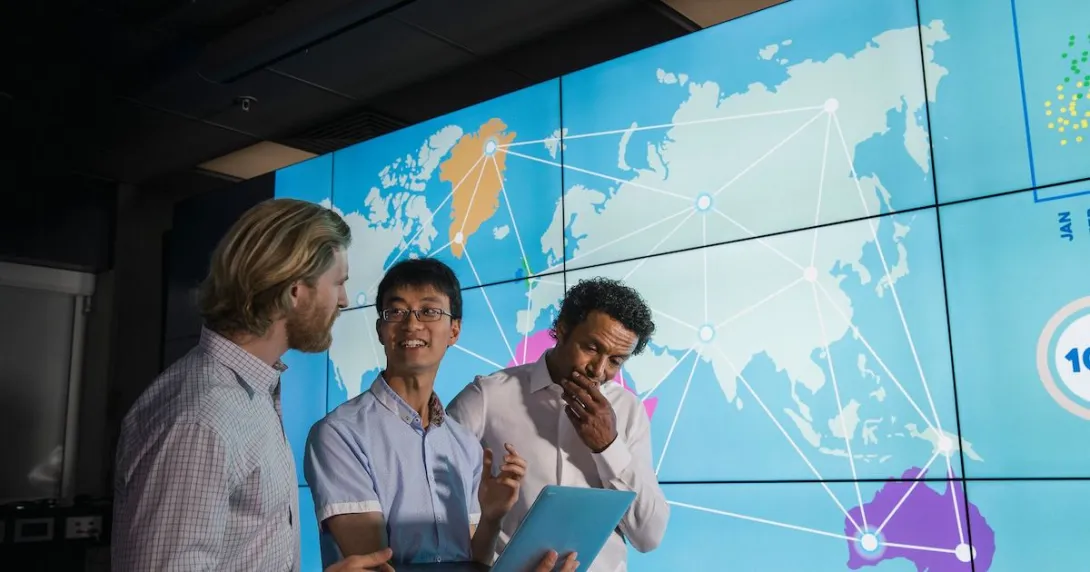 
  Three people standing in front of a board showing the world with lines connecting countries
